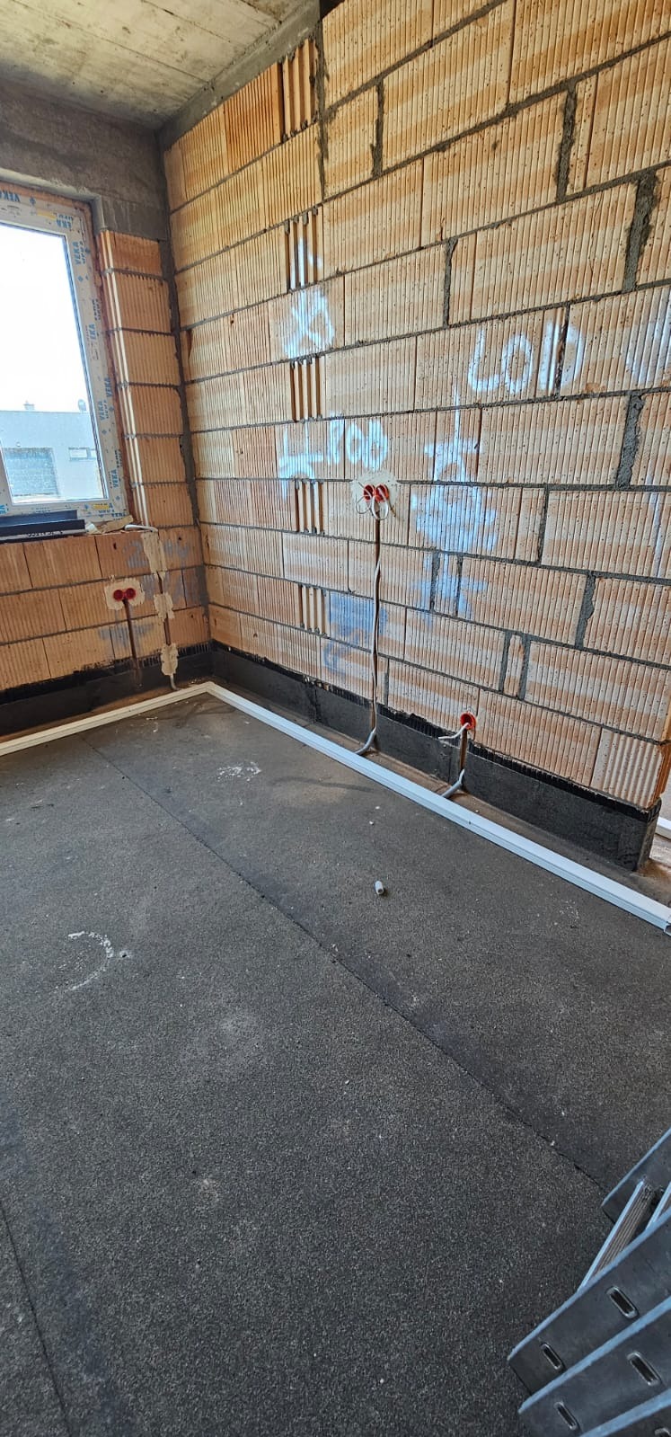 Surowy stan: Murowane ściany z widocznymi przewodami elektrycznymi i puszkami w ścianie, podłoga z izolacją, okno w rogu pomieszczenia. Wnętrze w trakcie prac.