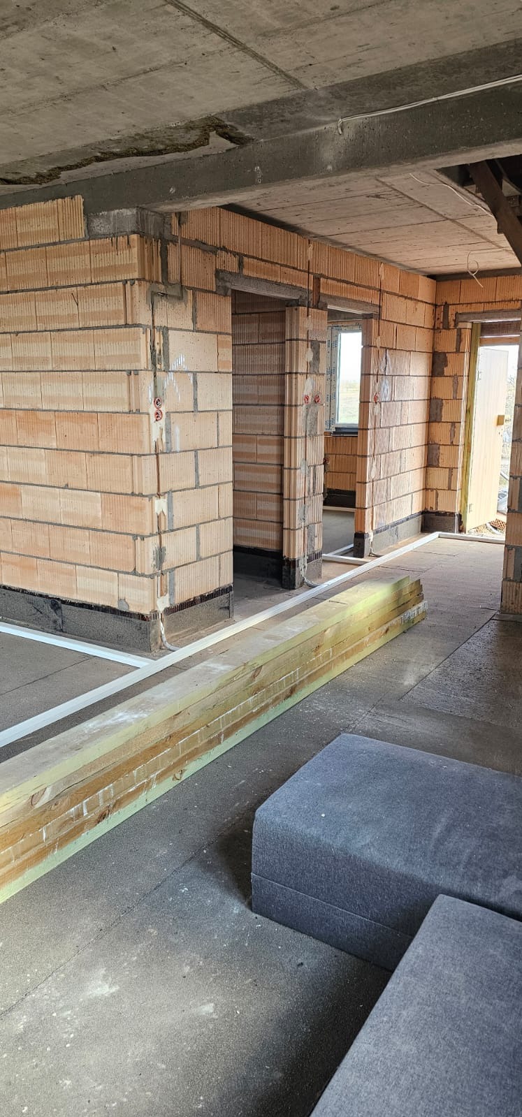 Surowy stan wnętrza budynku z ceglanymi ścianami i widocznymi przewodami elektrycznymi. Widoczne otwory drzwiowe i okienne. Na podłodze leżą deski.