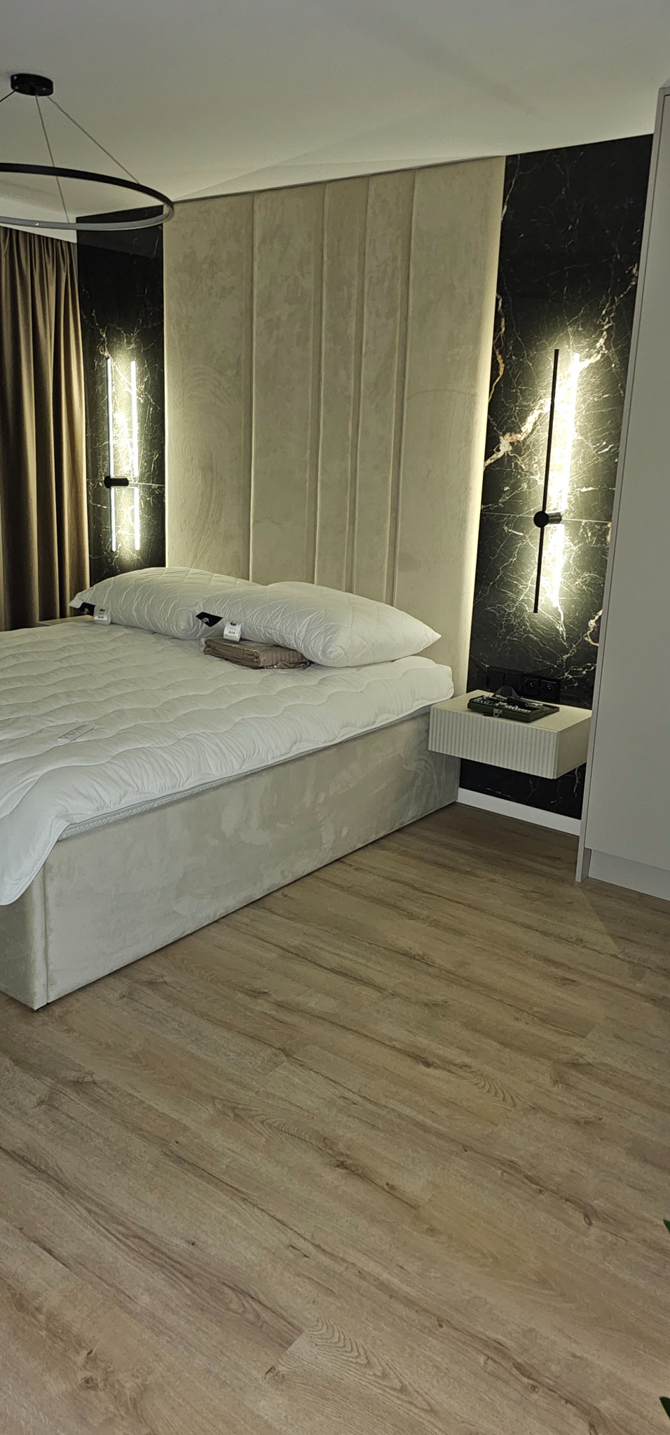 Nowoczesna sypialnia z podświetlanym panelem dekoracyjnym imitującym marmur i minimalistycznym oświetleniem liniowym przy łóżku z wezgłowiem.