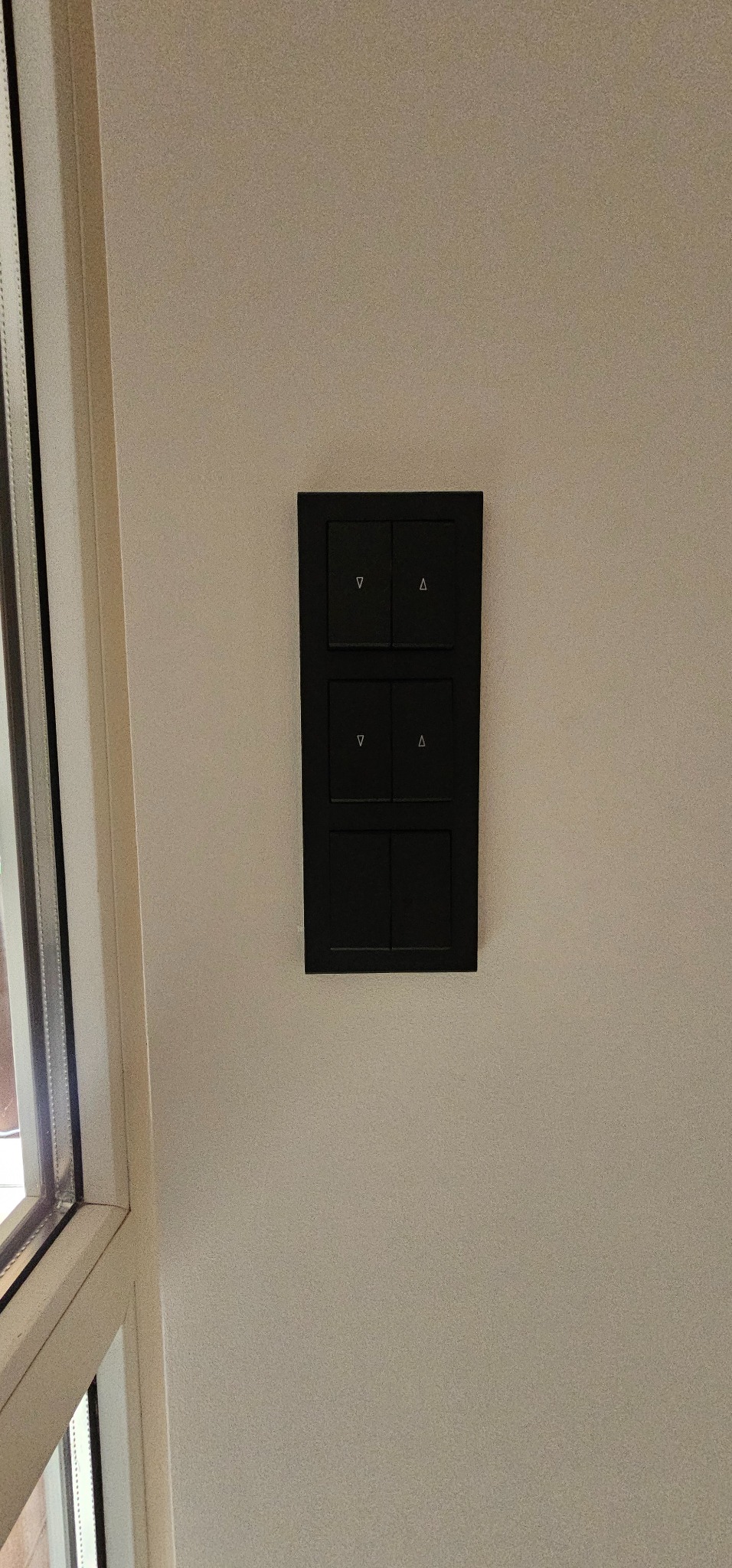 Czarny panel sterowania oświetleniem, zamontowany na kremowej ścianie, z widocznym fragmentem ramy okiennej po lewej stronie. Minimalistyczny design.