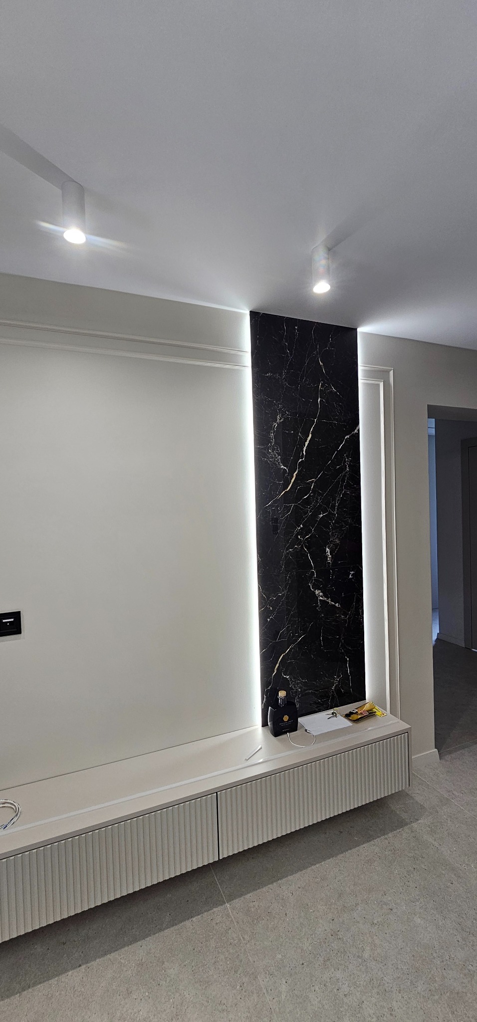 Nowoczesny salon z podświetlanym panelem z czarnego marmuru i białymi szafkami RTV. Dwa punktowe światła sufitowe oświetlają wnętrze. Minimalistyczny design.