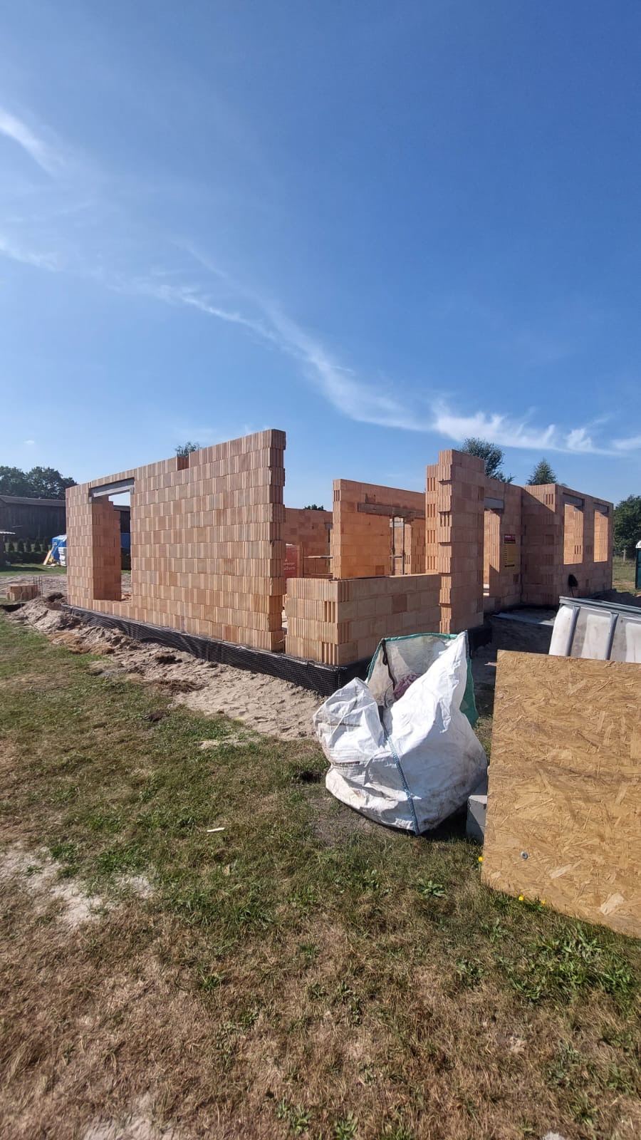 Wznoszone ściany domu z czerwonej cegły ceramicznej na fundamencie z widoczną izolacją, w tle błękitne niebo, na pierwszym planie biały worek budowlany i płyta OSB.