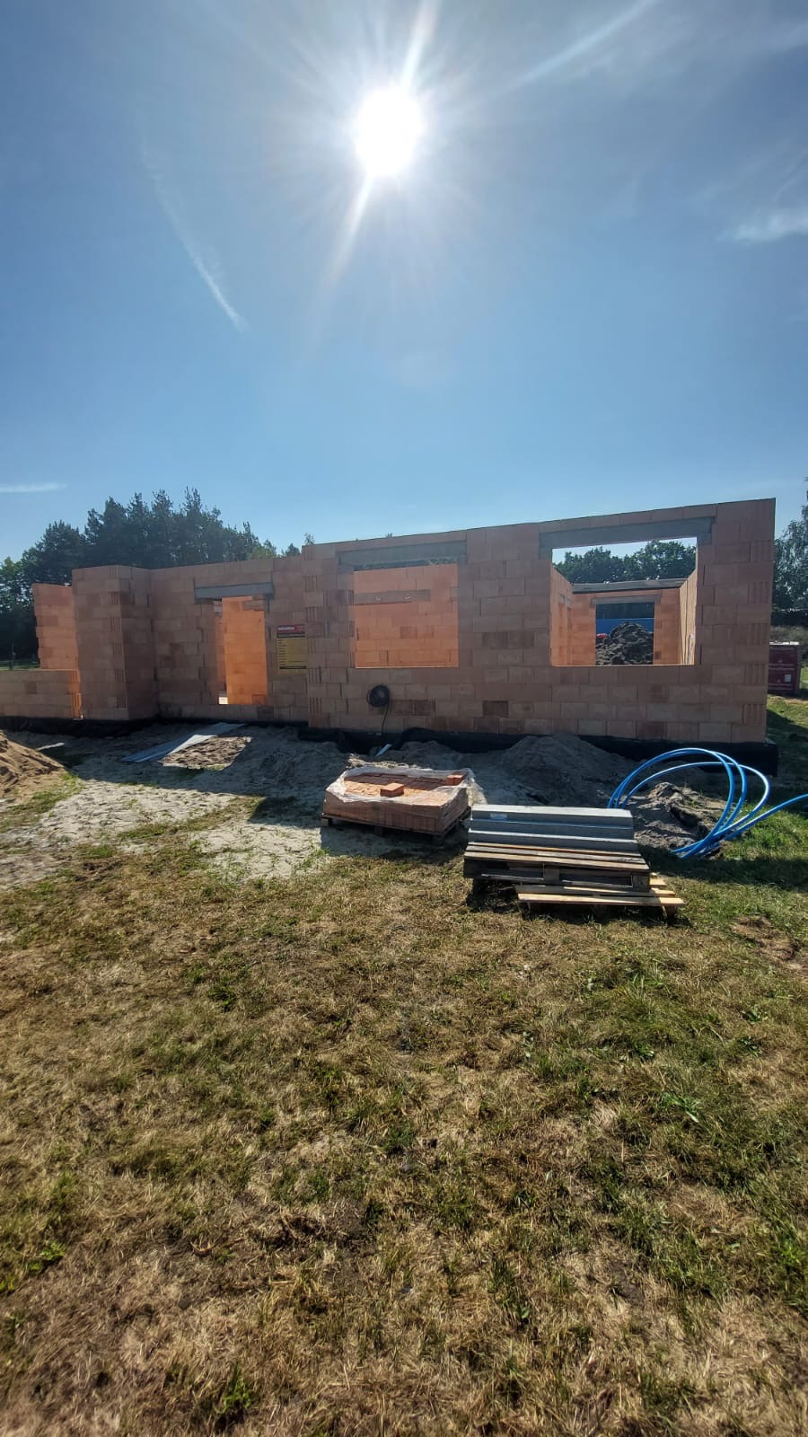 Wznoszone ściany z bloczków ceramicznych na budowie domu jednorodzinnego w słoneczny dzień, widoczne okna i drzwi w stanie surowym, paleta z bloczkami obok