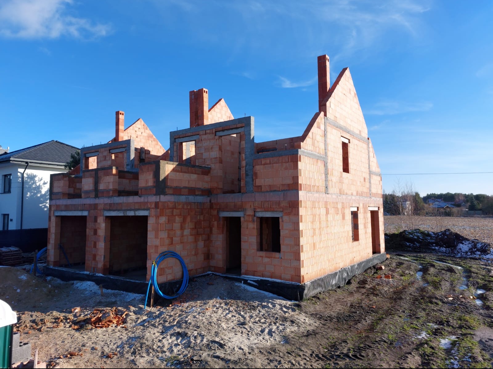 Wznoszenie dwukondygnacyjnego budynku z czerwonej cegły ceramicznej, widoczne otwory okienne i kominy w stanie surowym otwarte na błękitne niebo, na pierwszym planie piasek i niebieski wąż budowlany.