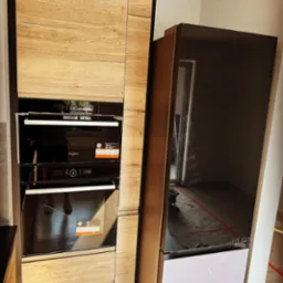 Nowoczesna zabudowa kuchenna z drewnianymi frontami, w której umieszczono piekarnik i mikrofalówkę marki Whirlpool, obok stoi czarna lodówka z białym dolnym panelem, widoczne odbicie w lodówce.