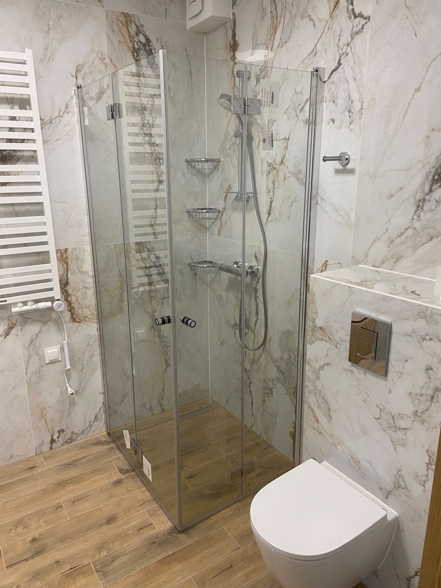Nowoczesna kabina prysznicowa z transparentnymi szklanymi ściankami, zamontowana w łazience wykończonej marmurowymi płytkami, z drewnopodobną podłogą i wiszącą toaletą.