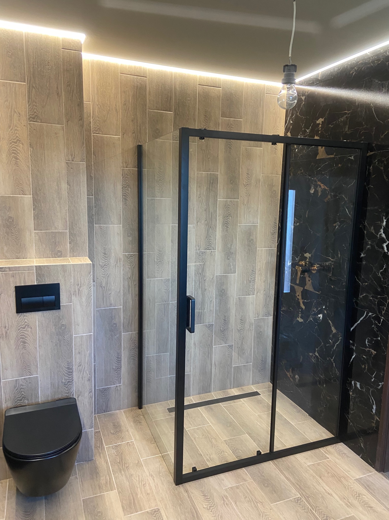 Nowoczesna łazienka z czarną kabiną prysznicową typu walk-in, podświetleniem LED podsufitowym, płytkami imitującymi drewno i wiszącą toaletą.