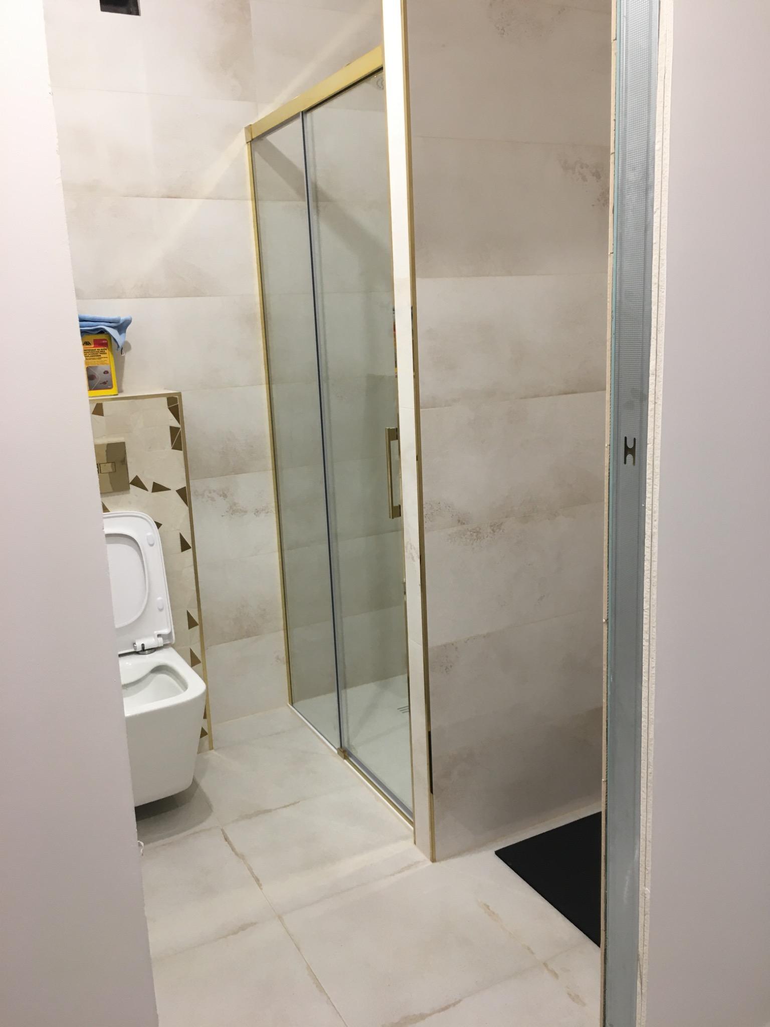 Wykończona łazienka z kabiną prysznicową w złotej ramie, toaletą wiszącą i płytkami imitującymi marmur, widok zza ściany z profilem metalowym.