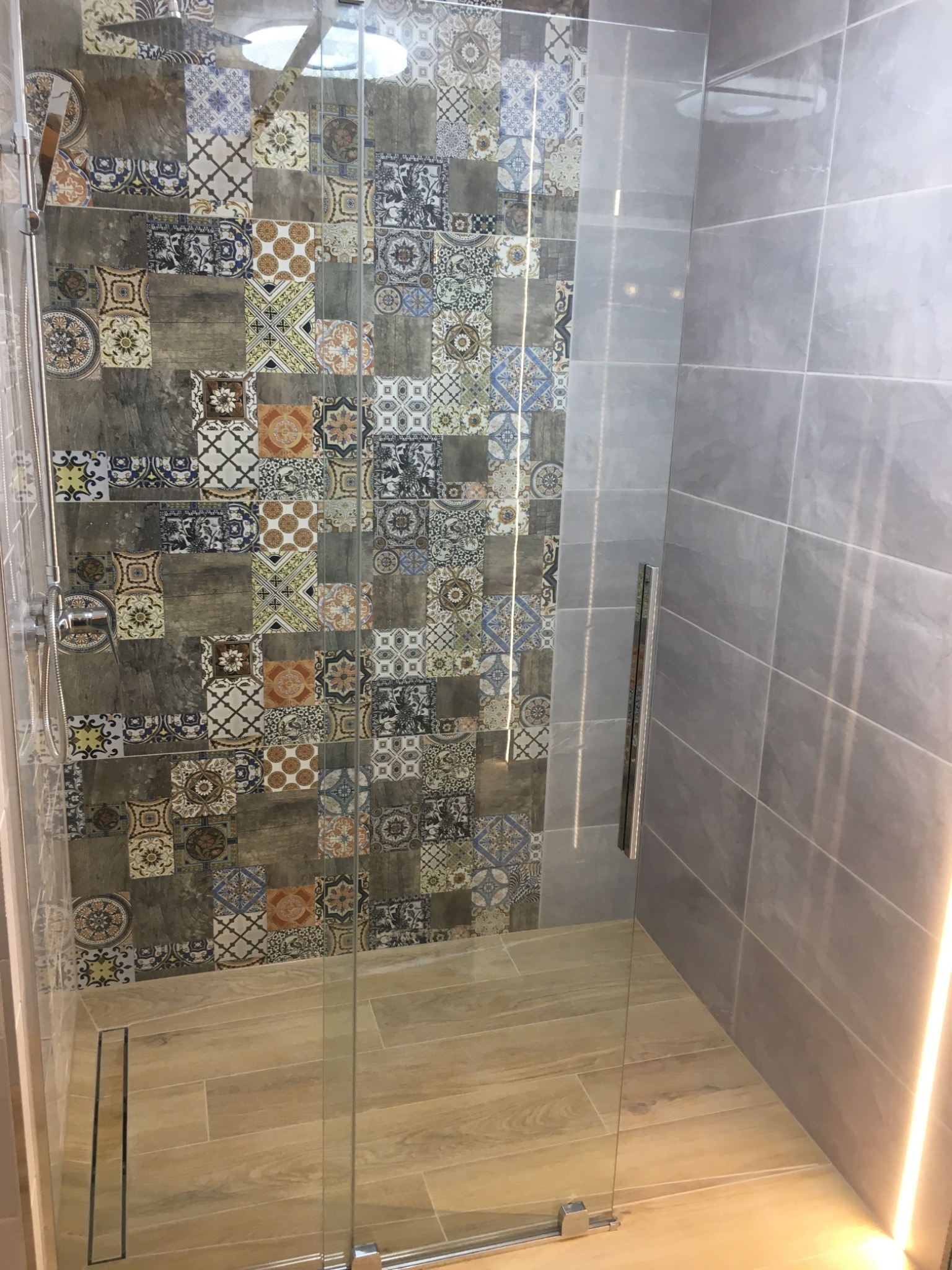 Nowoczesna kabina prysznicowa z mozaikową ścianą i szklanymi drzwiami, widoczny odpływ liniowy w podłodze wyłożonej płytkami imitującymi drewno.