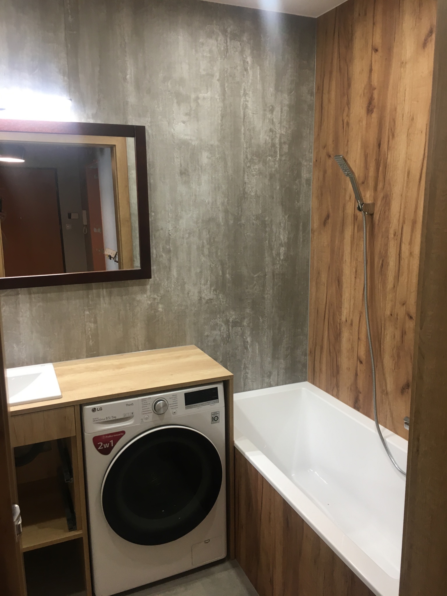 Nowoczesna łazienka z betonową ścianą, drewnianymi panelami, wanną i pralką LG zabudowaną pod umywalką.