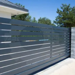 Nowoczesna, przesuwna brama wjazdowa z poziomymi panelami w kolorze grafitowym, zintegrowana z ogrodzeniem panelowym i słupkami z szarego betonu architektonicznego, na tle domu z szarą dachówką...