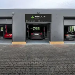 Elewacja warsztatu samochodowego z trzema stanowiskami, w środkowym samochod marki Tesla na podnośniku, nad wjazdem szyld z nazwą warsztatu i danymi kontaktowymi.