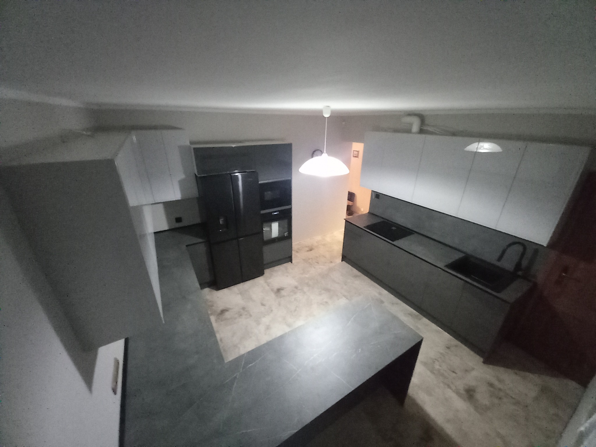 Nowoczesna kuchnia z szarymi frontami, czarnym sprzętem AGD i wyspą kuchenną z blatem imitującym marmur, oświetlona lampą wiszącą.