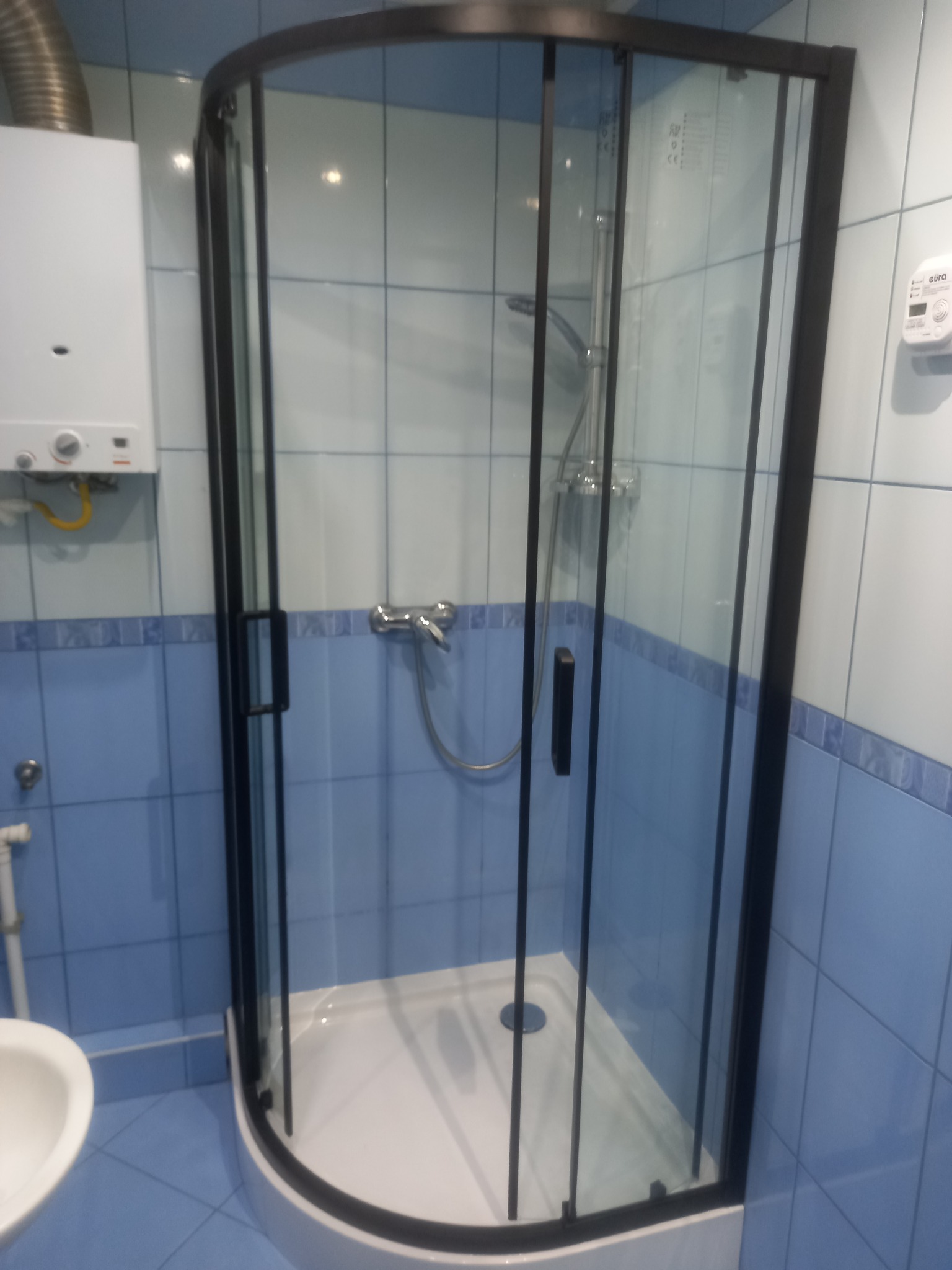 Kabina prysznicowa narożna z czarnymi profilami i brodzikiem, na tle niebieskich płytek, widoczny bojler i umywalka.