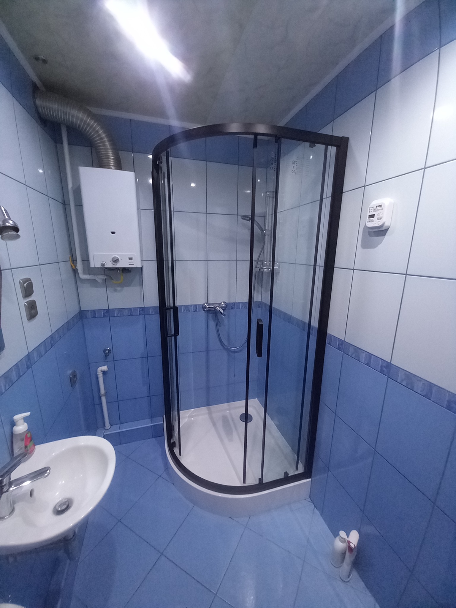 Narożna kabina prysznicowa z czarną ramą w niebiesko-białej łazience z płytkami, widoczny bojler gazowy i umywalka.