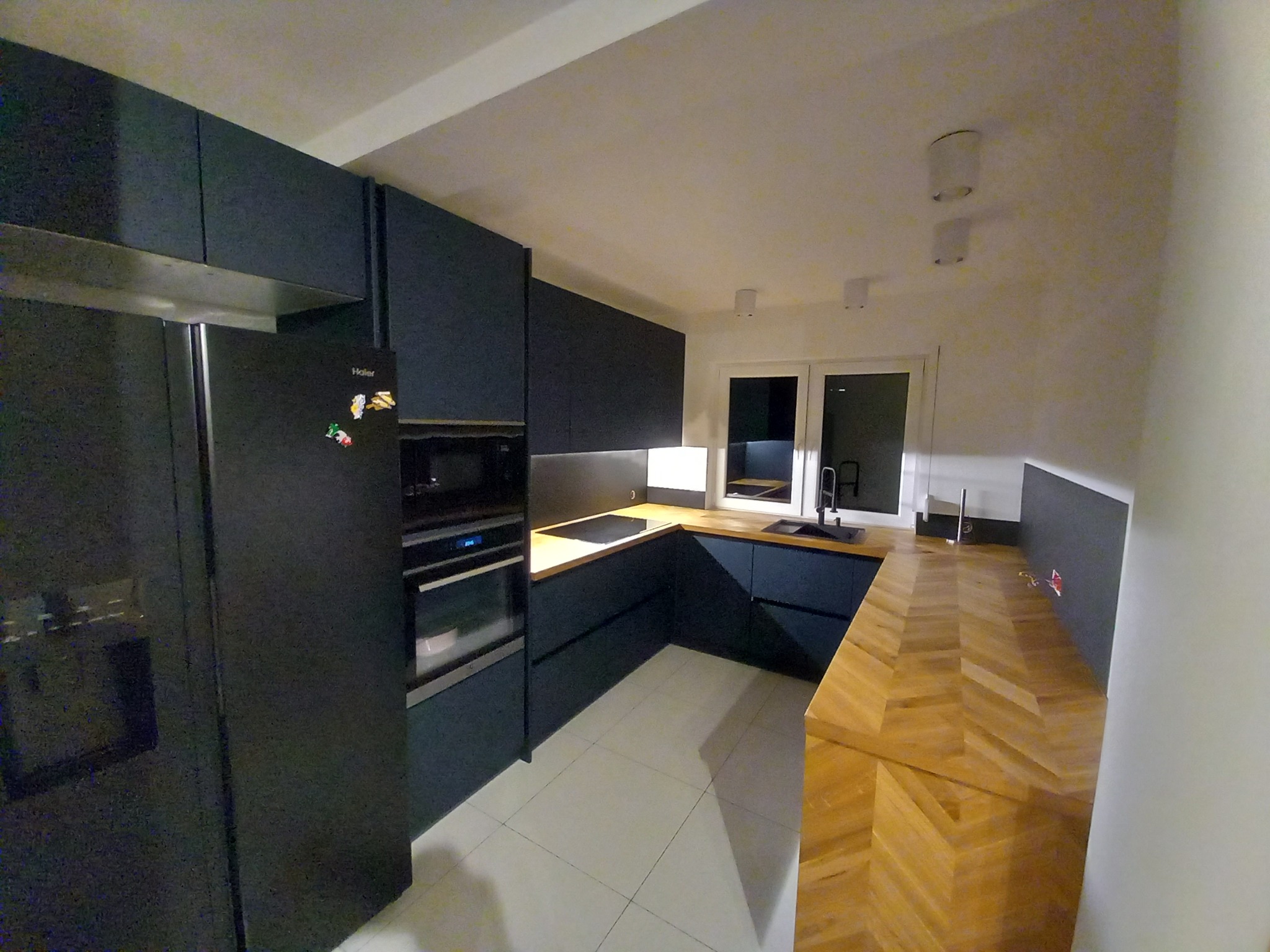 Nowoczesna kuchnia z ciemnymi, matowymi szafkami, drewnianym blatem ułożonym we wzór jodełki i białymi płytkami na podłodze, widziana z perspektywy szerokokątnej.