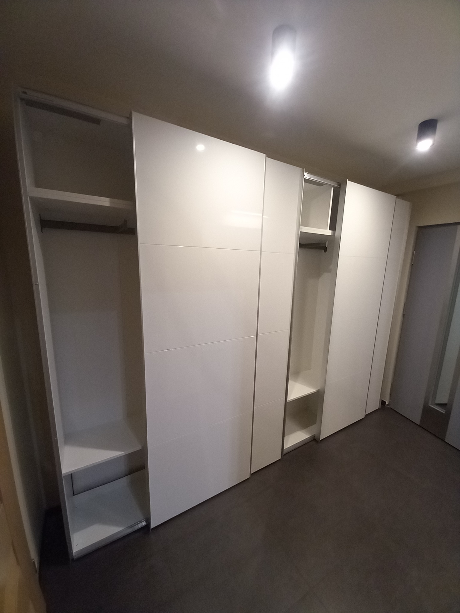 Biała szafa wnękowa z przesuwnymi drzwiami, widoczne półki i drążek na ubrania, nowoczesny design, ciemna podłoga.