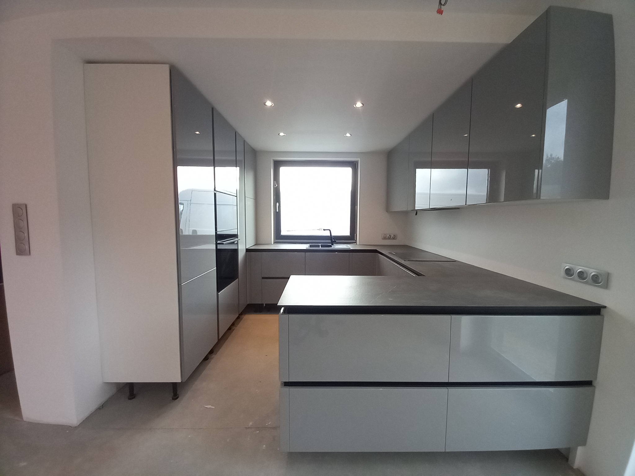 Nowoczesna, szara kuchnia z wyspą, fronty szafek z połyskiem, widoczne gniazdka elektryczne na ścianie, okno z widokiem na zewnątrz.