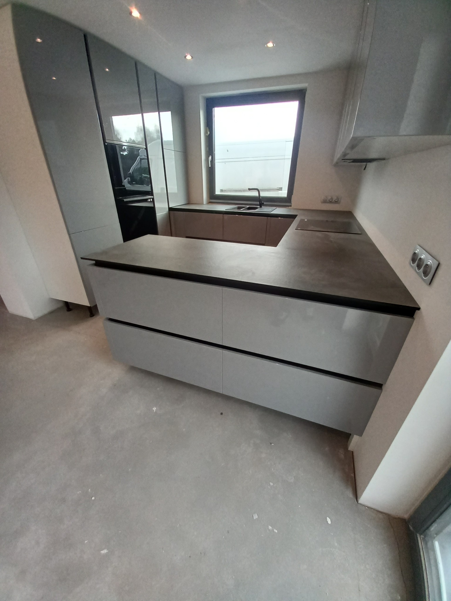 Nowoczesna kuchnia z szarymi, lakierowanymi frontami, ciemnym blatem i oknem z widokiem na zewnątrz, jeszcze przed pełnym wykończeniem podłogi.