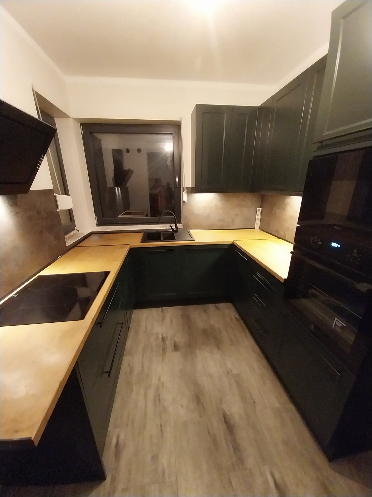 Nowoczesna, ciemnozielona zabudowa kuchenna z drewnianym blatem i czarną płytą indukcyjną w Rybniku, widziana z perspektywy podłogi.