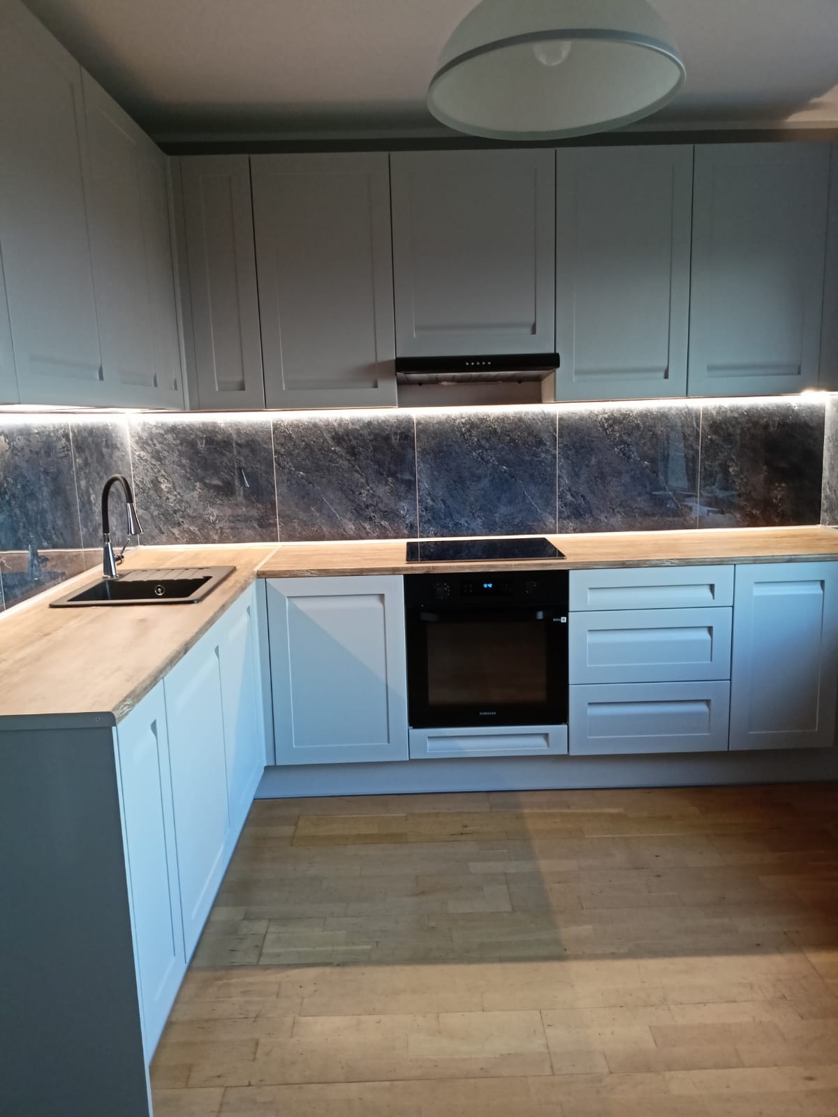 Jasnoszara zabudowa kuchenna z drewnianym blatem, czarnym zlewem i płytą indukcyjną, oświetlenie LED pod szafkami górnymi, widok na parkiet.