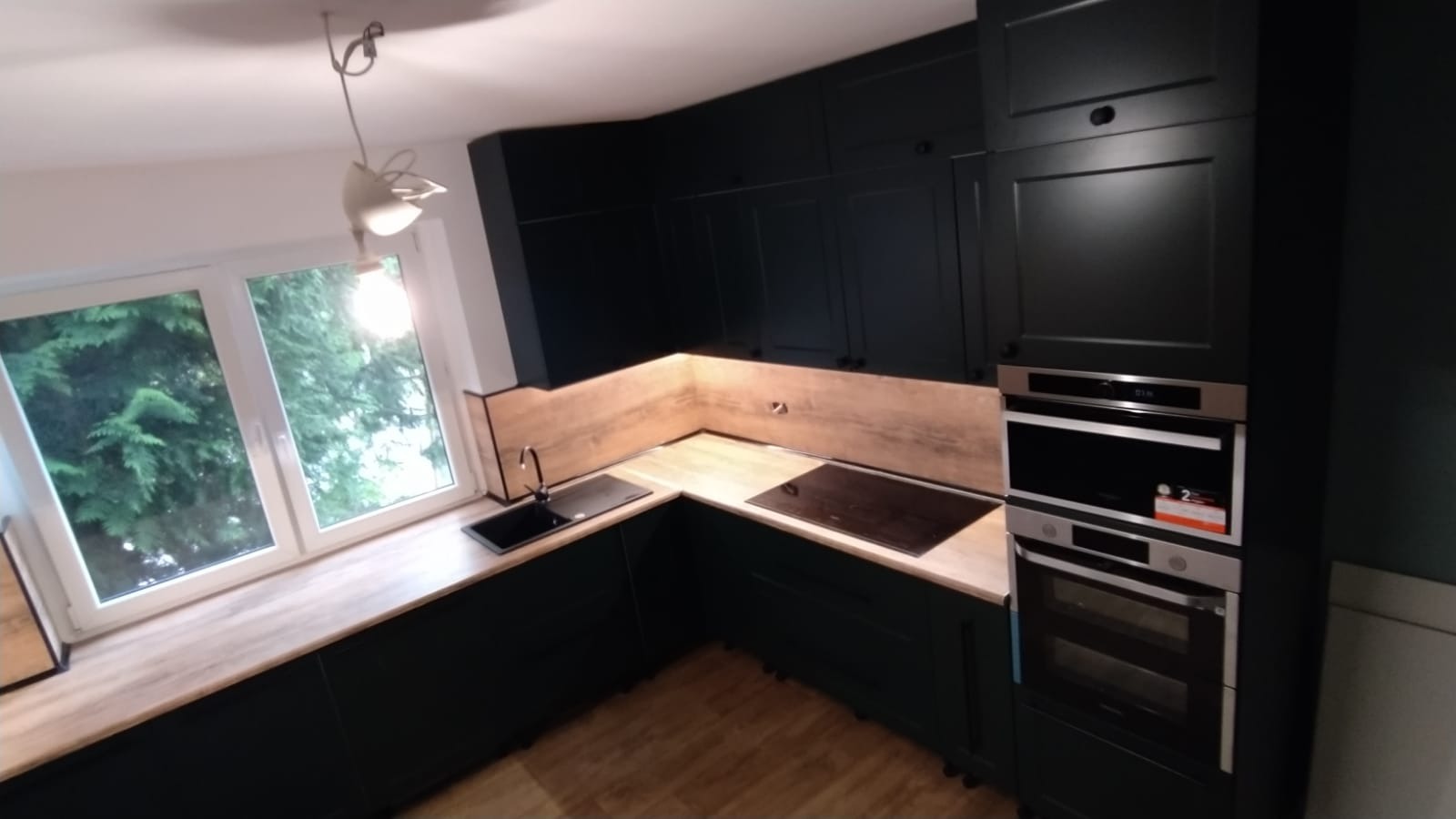 Ciemnozielona kuchnia na wymiar z drewnianym blatem, czarnym zlewem i płytą indukcyjną, oświetlona lampą w kształcie ptaka, widok z okna na zieleń.