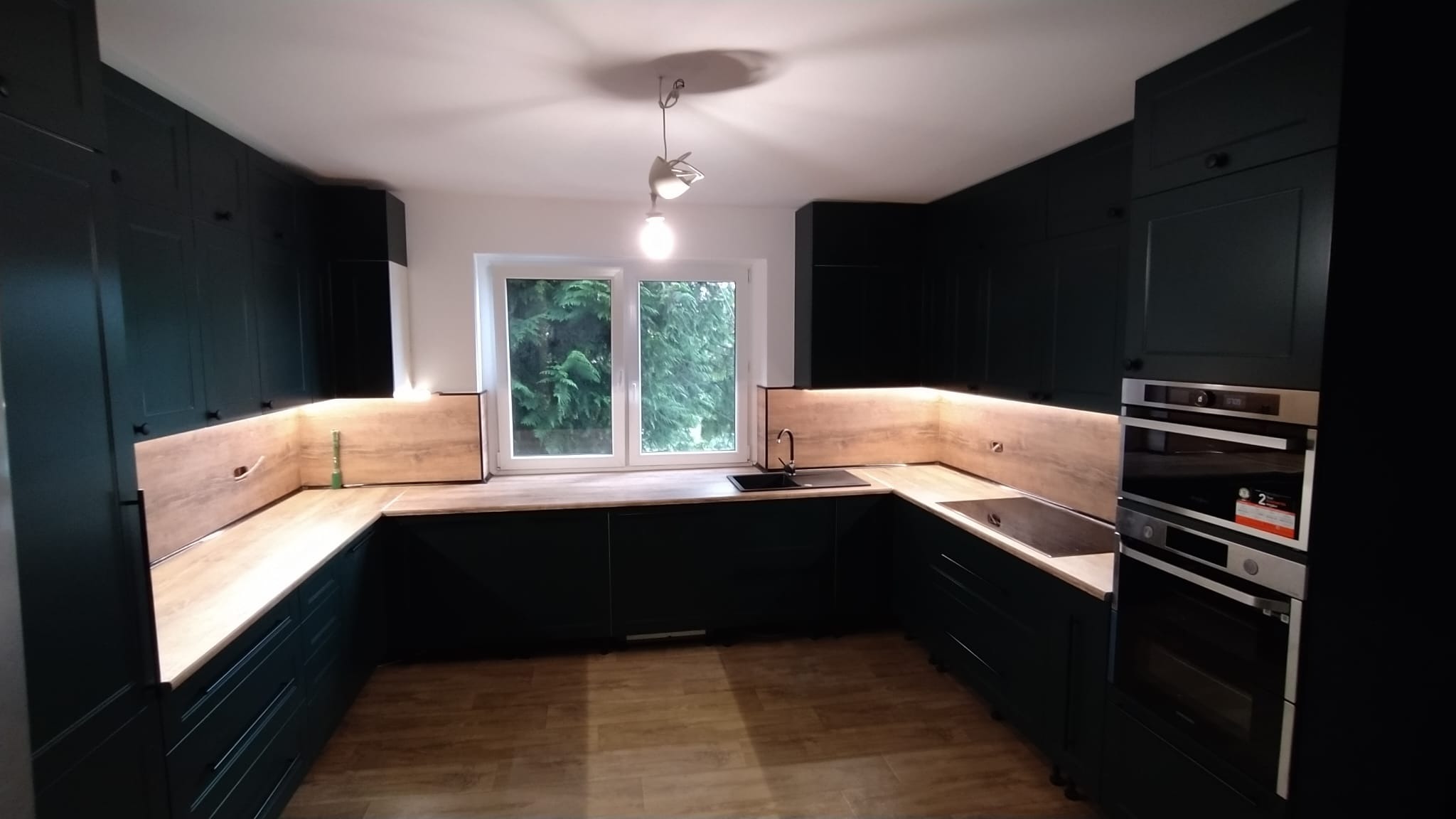 Nowoczesna, ciemnozielona kuchnia z drewnianym blatem i oświetleniem LED pod szafkami wiszącymi, widok na okno z zielenią na zewnątrz.