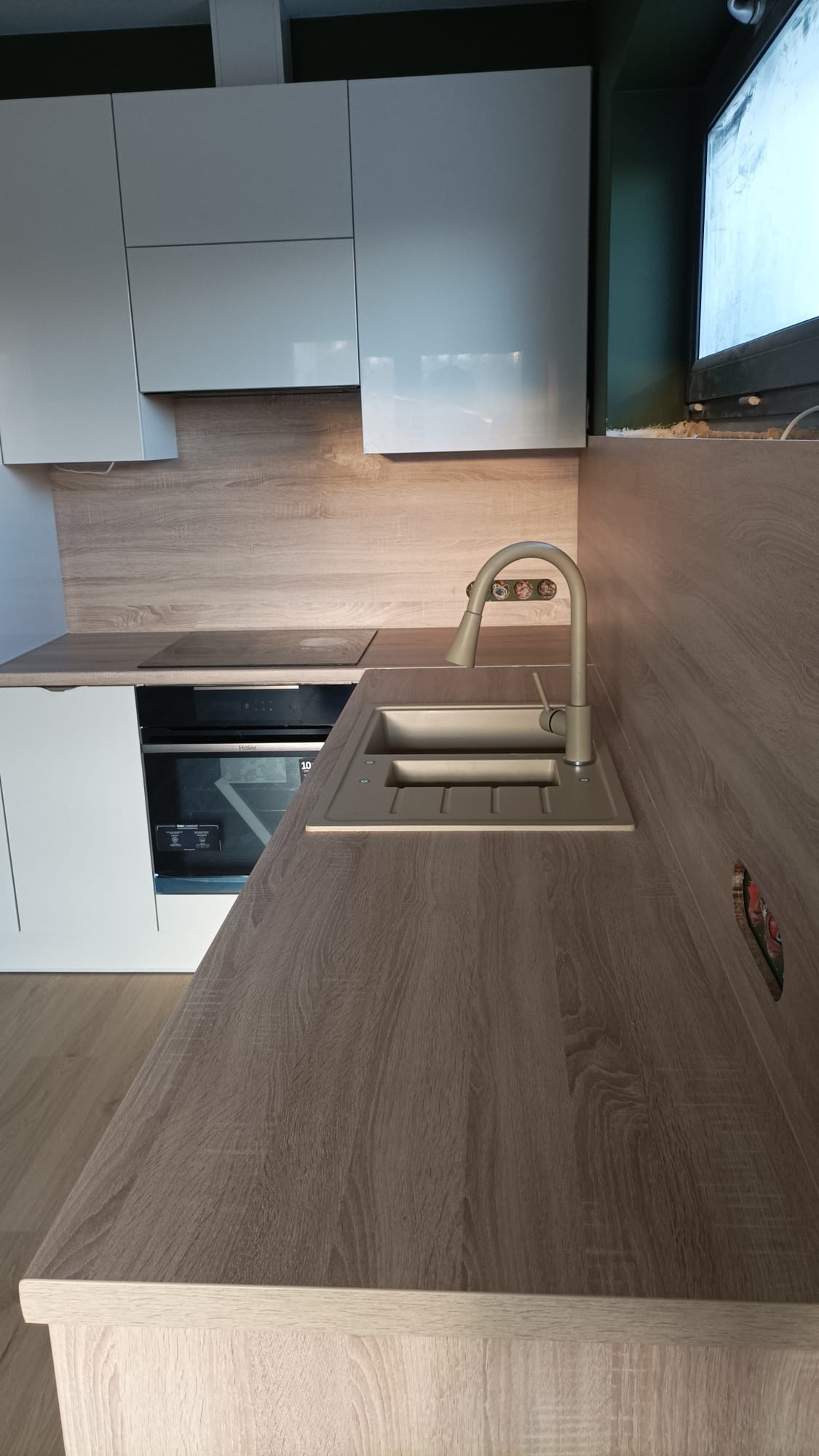 Nowoczesna kuchnia z jasnymi, lakierowanymi szafkami górnymi, blatem imitującym drewno, zlewem w kolorze blatu i płytą indukcyjną. Widoczny fragment okna z ciemnozieloną ramą.