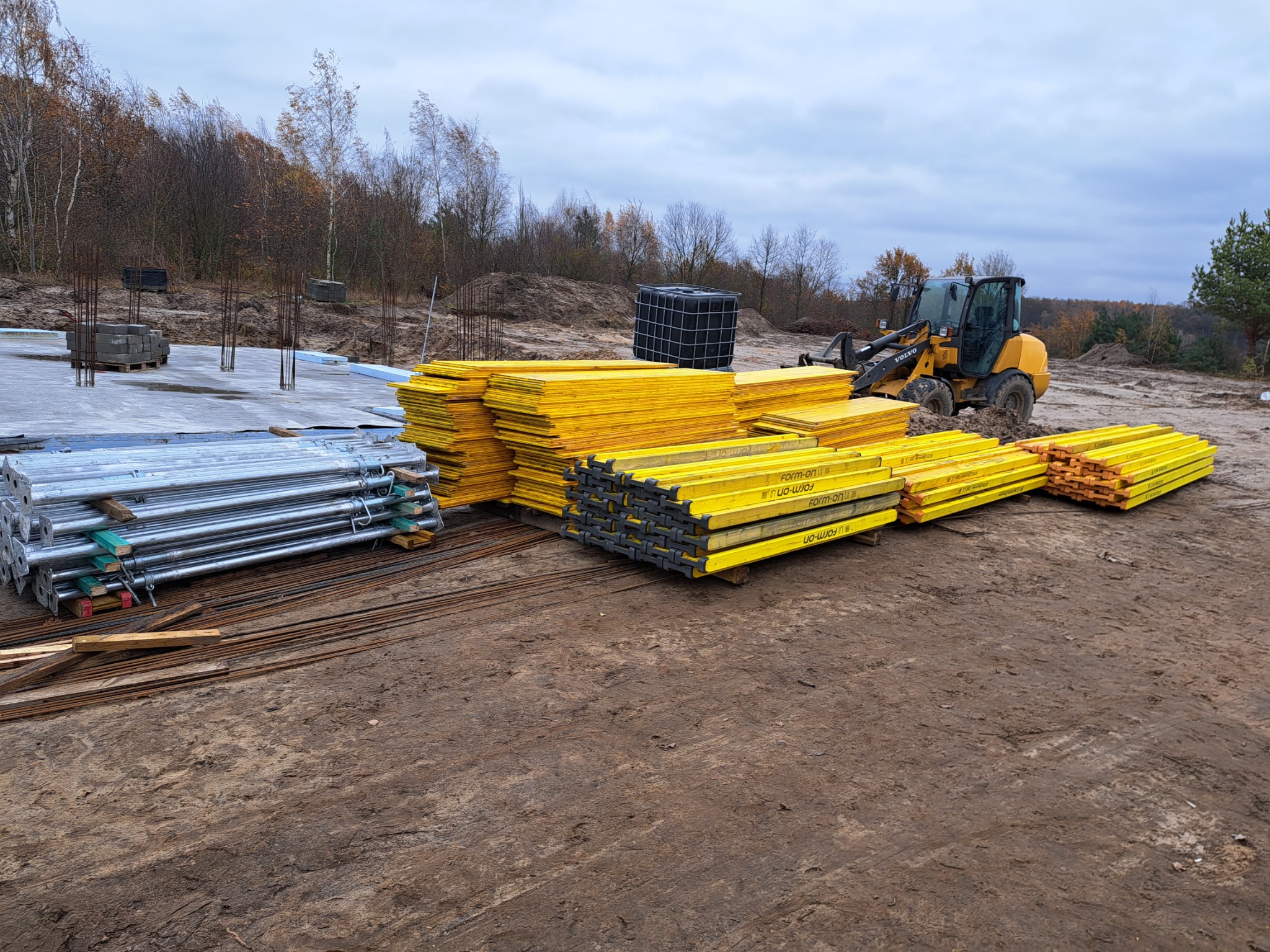 Stosy żółtych szalunków budowlanych i metalowych podpór na placu budowy z widocznym fundamentem i maszyną budowlaną w tle.