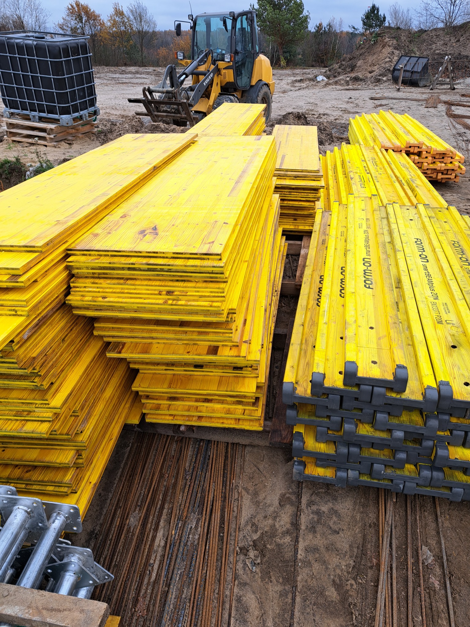 Stosy żółtych szalunków budowlanych Form-on z czarnymi elementami, ułożone na placu budowy z widocznymi prętami zbrojeniowymi i ładowarką Volvo w tle.