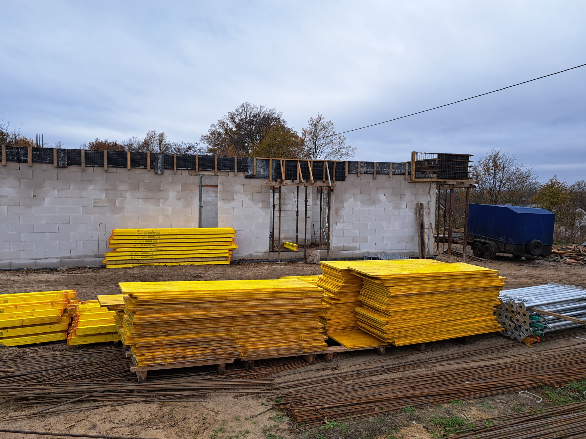 Budowa domu w trakcie: ściany z betonu komórkowego, szalunki zalane betonem, żółte deski szalunkowe ułożone na ziemi, zbrojenia stalowe, przyczepa w tle.