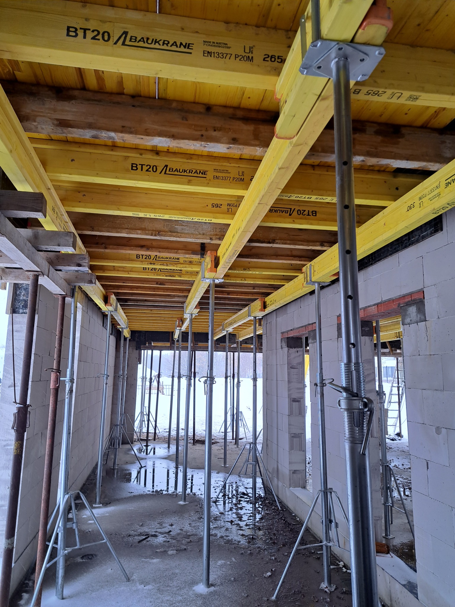 Wnętrze budynku w trakcie budowy: widoczne białe ściany z betonu komórkowego, podparte żółtymi belkami stropowymi z widocznymi oznaczeniami 'BT20/BAUKRANE' i metalowymi stemplami budowlanymi,...