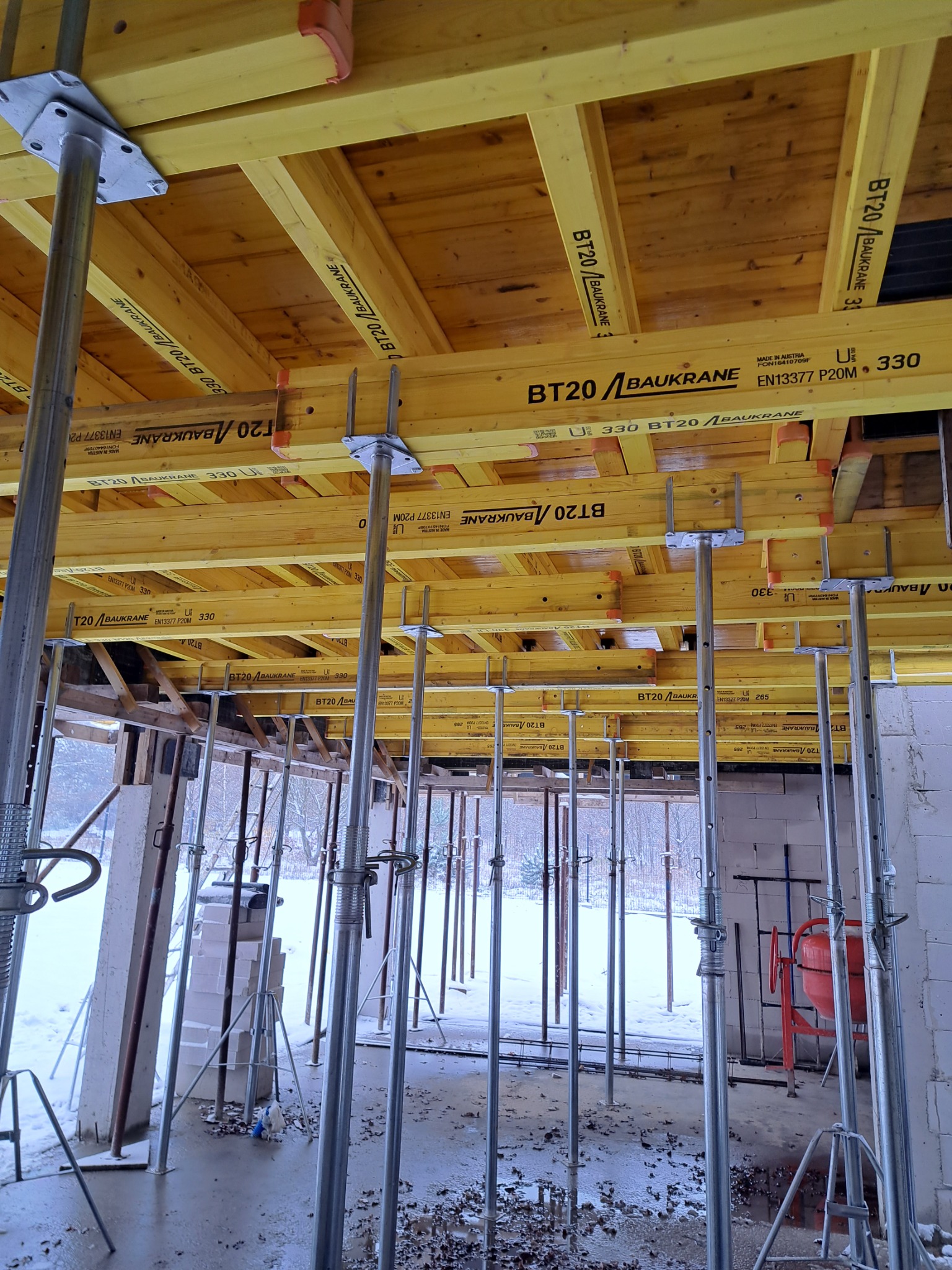 Wnętrze budynku w trakcie budowy z widocznymi żółtymi belkami stropowymi podpartymi metalowymi stemplami budowlanymi, na zewnątrz widok na ośnieżony teren.