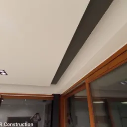 Wykończony sufit z oświetleniem punktowym i dekoracyjnym pasem w kolorze antracytowym, widok z perspektywy pomieszczenia z dużymi oknami przesuwnymi.