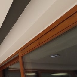 PerfektBud - Fragment sufitu z pomalowanymi pasami w kolorach szarym i białym, oraz drewniane obramowanie okna przesuwnego z widocznym odbiciem wnętrza pomieszczenia i oświetleniem.