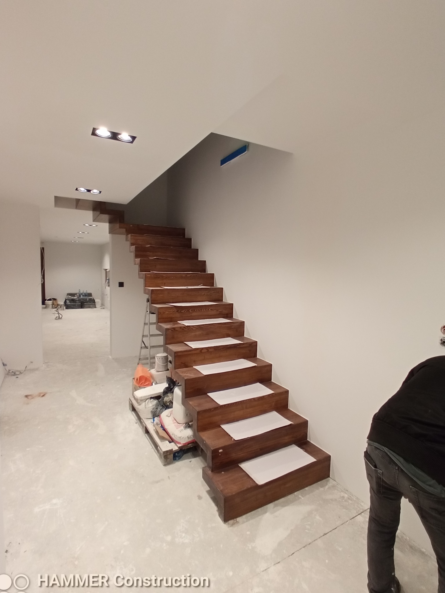 Drewniane schody z białymi zabezpieczeniami na stopniach podczas remontu, widoczne narzędzia i materiały budowlane na palecie, surowe ściany i oświetlenie punktowe w nowym domu.