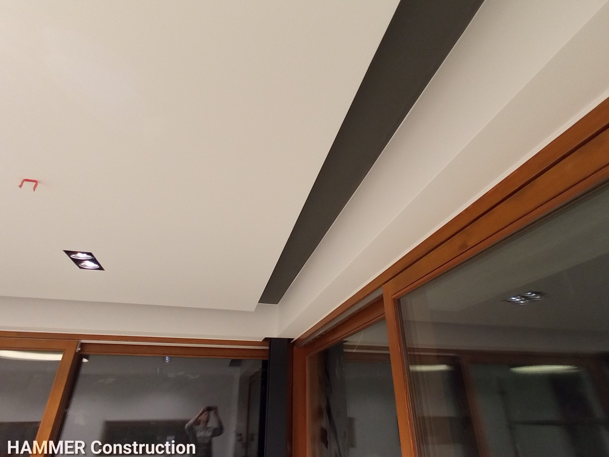 Wykończony sufit z oświetleniem punktowym i dekoracyjnym pasem w kolorze antracytowym, widok z perspektywy pomieszczenia z dużymi oknami przesuwnymi.