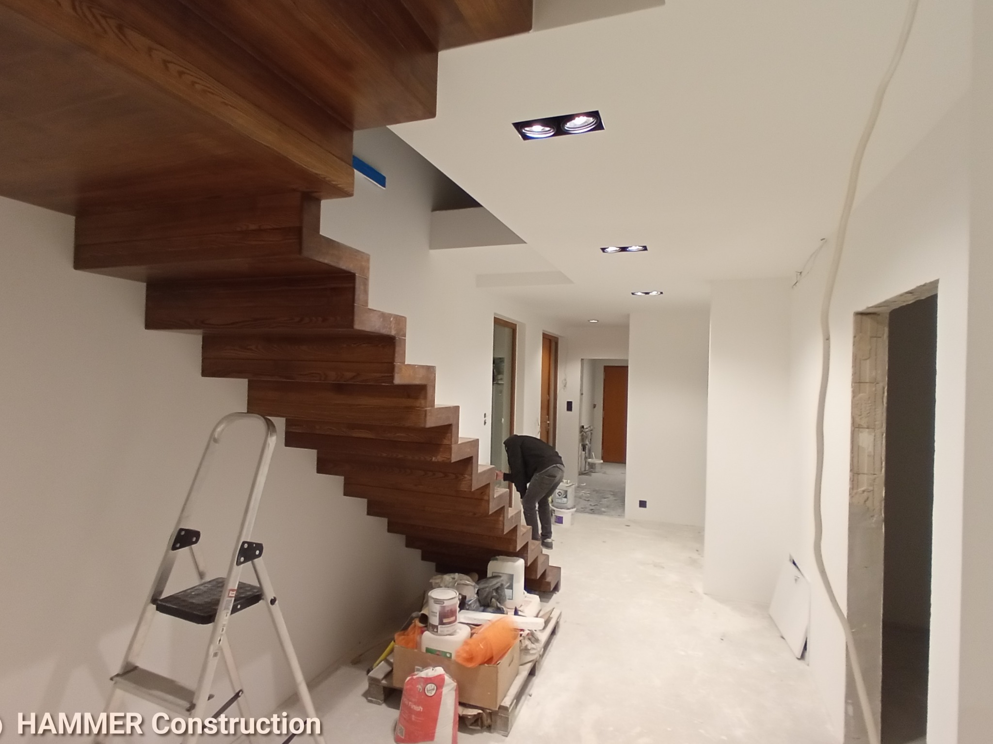 Montaż drewnianych schodów wspornikowych w nowoczesnym wnętrzu, widoczna drabina malarska i materiały budowlane, w tle osoba wykonująca prace wykończeniowe.