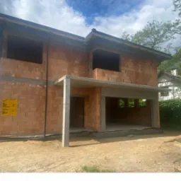 Dwukondygnacyjny dom murowany w stanie surowym otwartym, z widoczną konstrukcją dachu, otwory okienne bez ram, żółta tablica informacyjna na ścianie, ziemia wokół budynku.