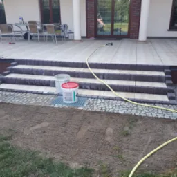 Wykończone kamienne schody wejściowe z jasnymi stopniami i ciemnymi obrzeżami prowadzące na taras z jasnych płytek, obok widoczna kostka brukowa i przygotowany teren pod trawnik, żółty wąż ogrodowy...