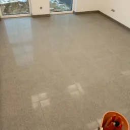 Wnętrze pomieszczenia z nową, jasną, gładką podłogą wyłożoną kwadratowymi płytkami. Widoczny kubeł z mopem w rogu, sugerujący świeżo zakończone prace porządkowe. Za oknami widoczny teren budowy.