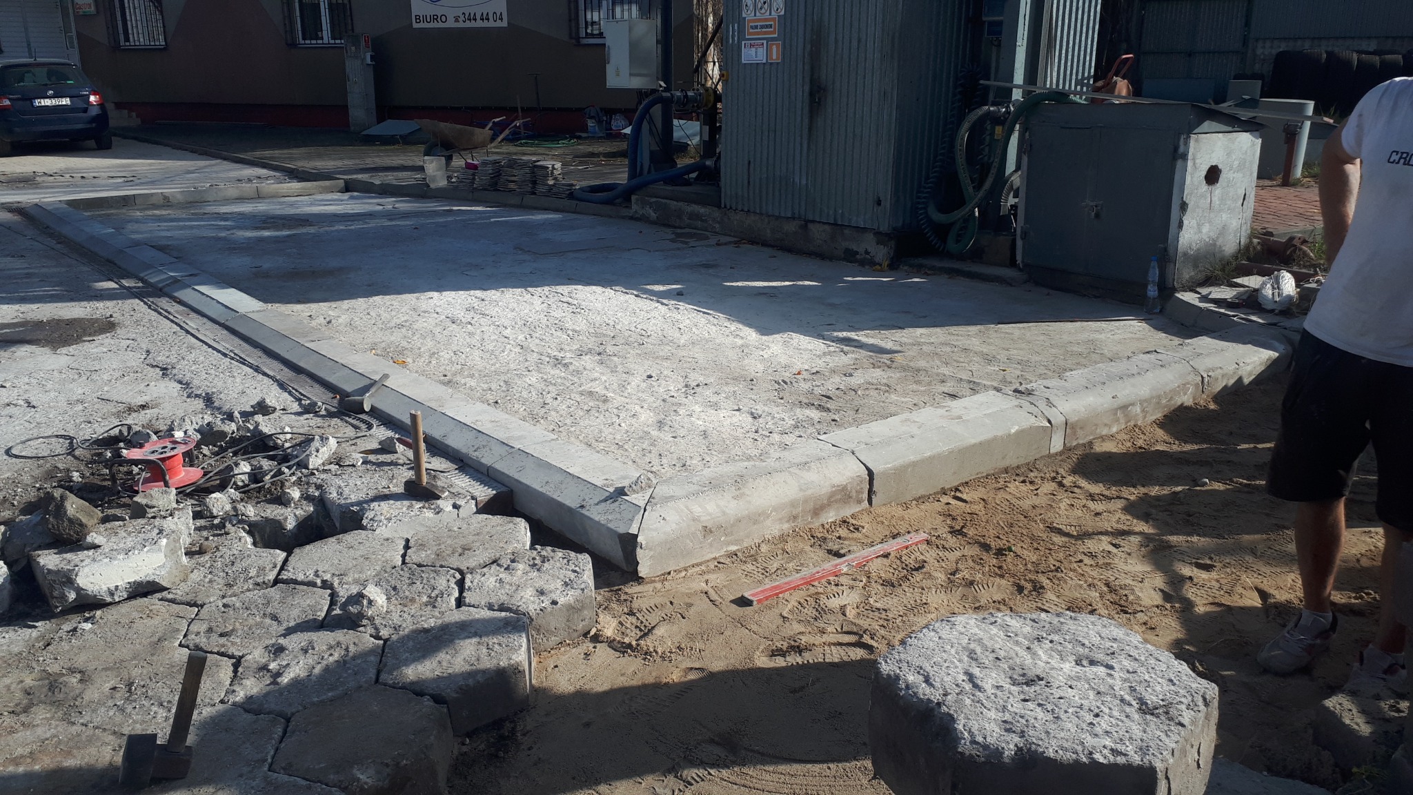 Przygotowany teren z obrzeżami betonowymi, podsypką piaskową i częściowo ułożoną kostką brukową w kształcie nieregularnych kamieni, widoczne narzędzia i akcesoria budowlane.