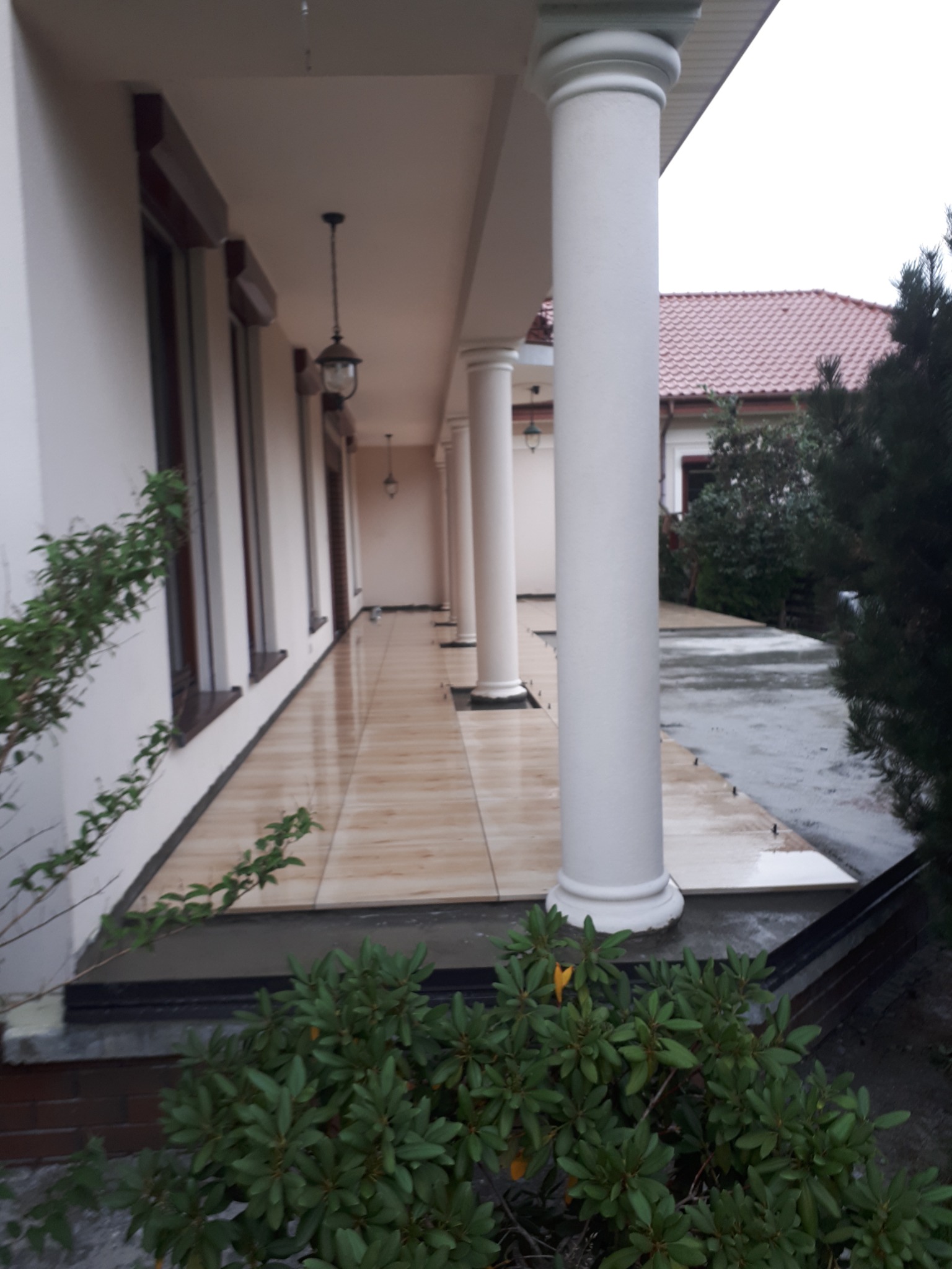 Zadaszone przejście z beżową, błyszczącą podłogą wyłożoną płytkami, wsparte na białych, okrągłych kolumnach, z wiszącymi lampami i widoczną zielenią w tle.