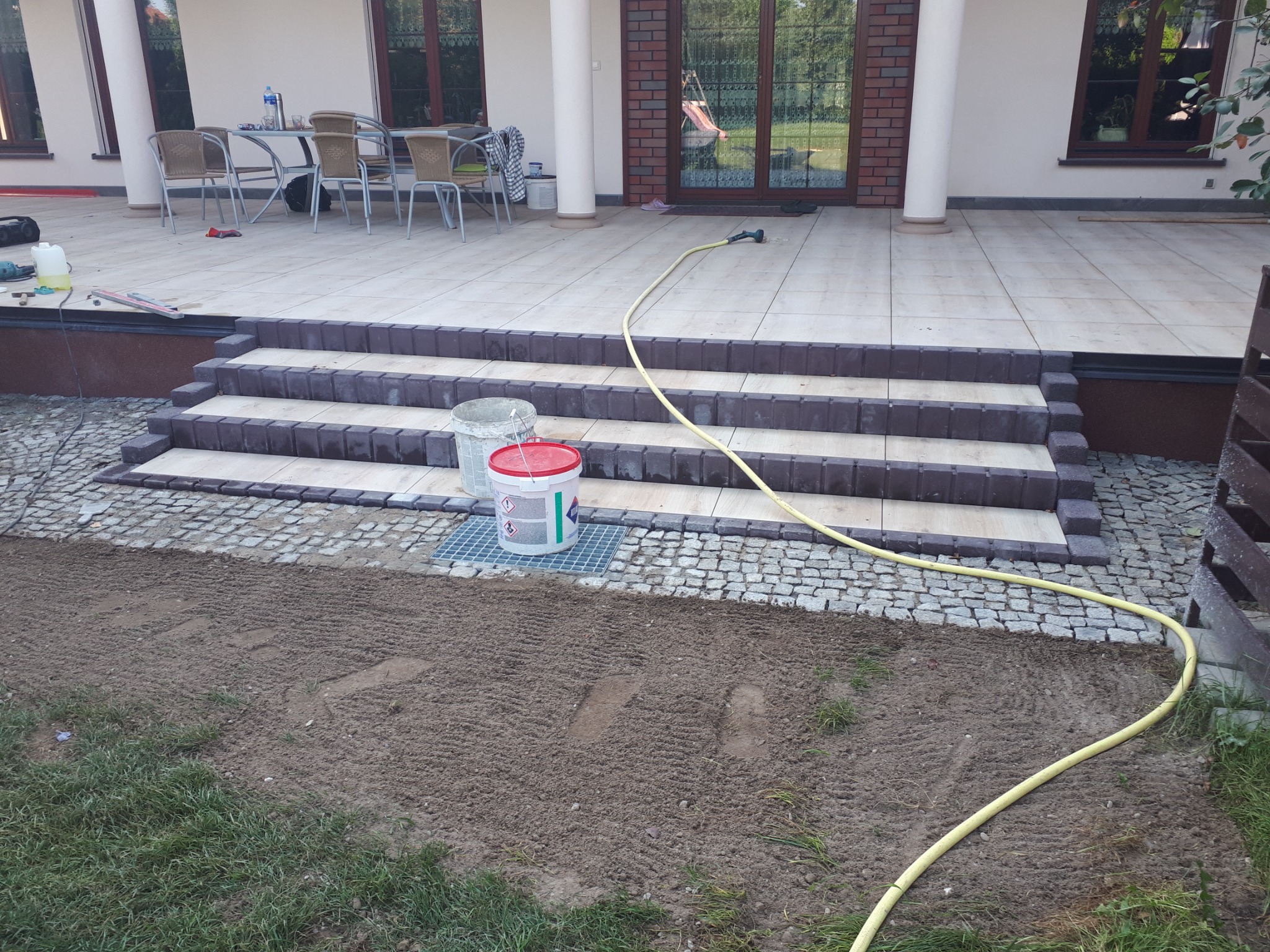 Wykończone kamienne schody wejściowe z jasnymi stopniami i ciemnymi obrzeżami prowadzące na taras z jasnych płytek, obok widoczna kostka brukowa i przygotowany teren pod trawnik, żółty wąż ogrodowy...