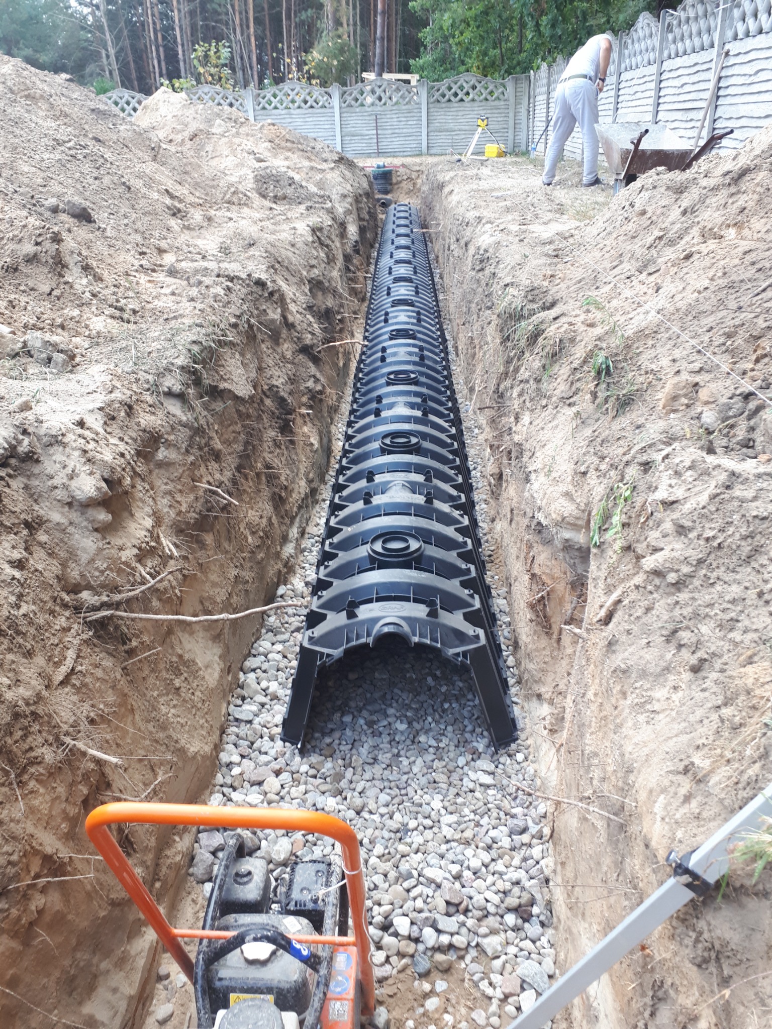 Drenaż liniowy z rur perforowanych ułożony w wykopie wypełnionym żwirem, widoczny zagęszczarka wibracyjna i pracownik z taczką w tle.