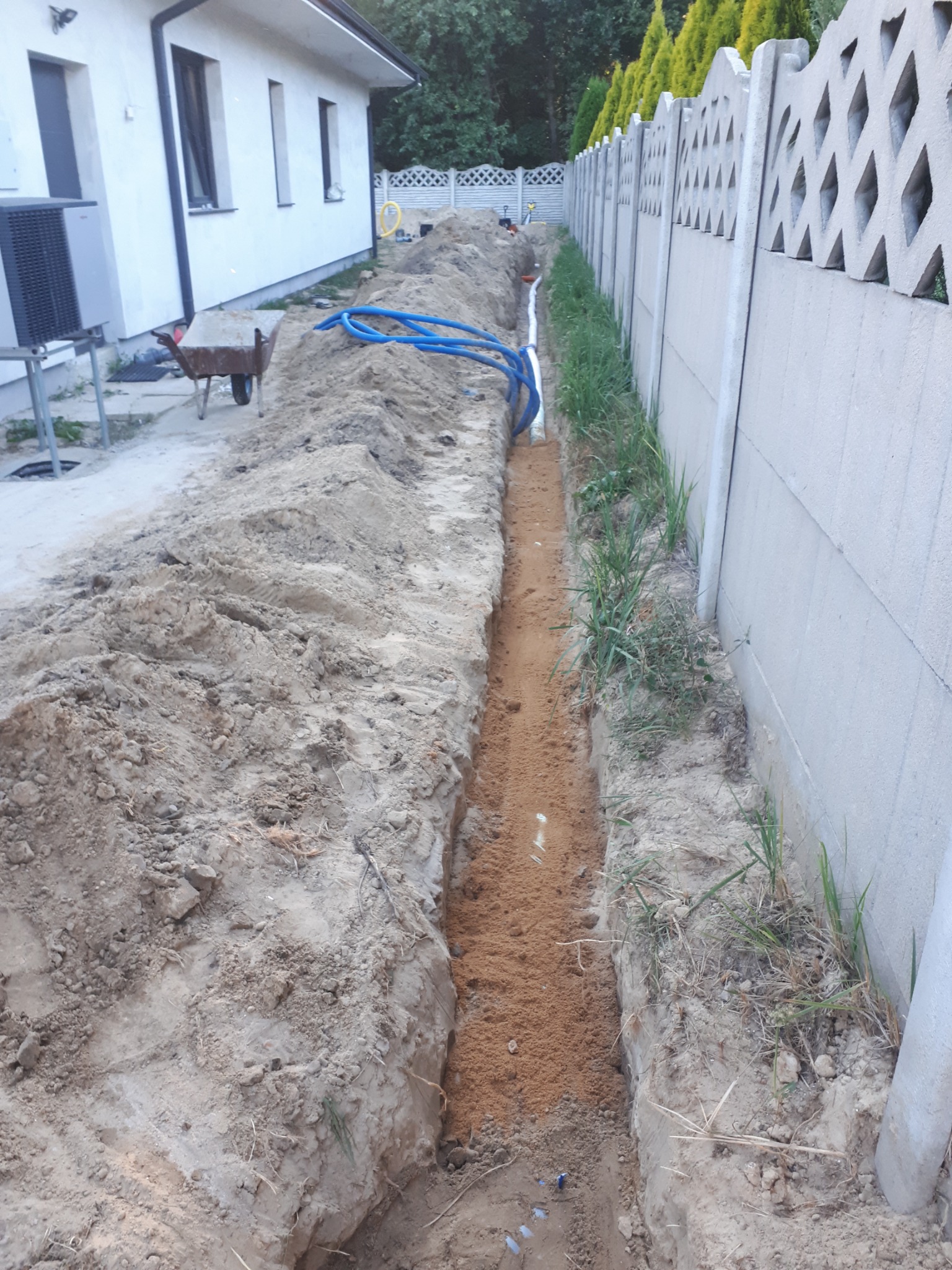 Wykop w ziemi wzdłuż fundamentu domu i betonowego ogrodzenia, z widocznymi niebieskimi rurami i warstwą piasku na dnie, przygotowany do instalacji.