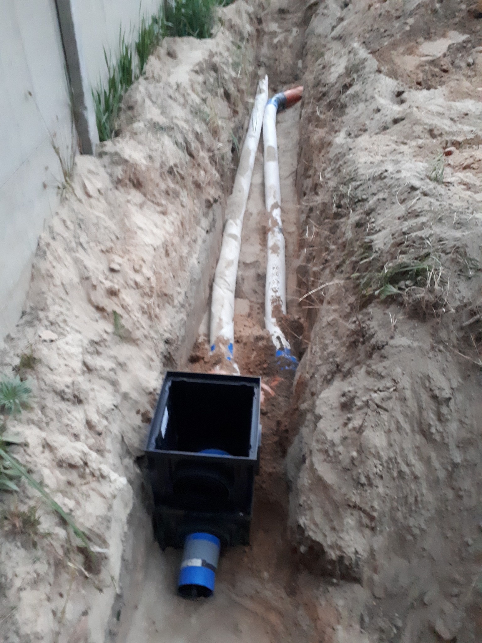 Wykop z ułożonymi rurami kanalizacyjnymi, częściowo owiniętymi izolacją, oraz czarną studzienką odpływową z niebieskim elementem łączącym.