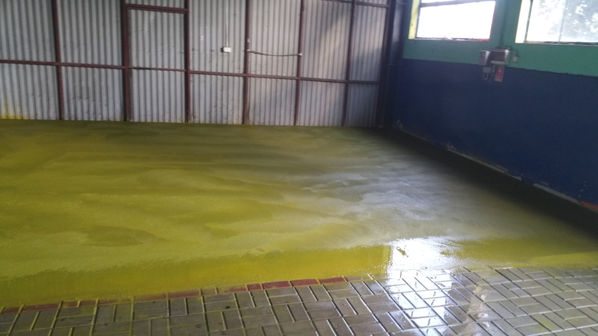 Żółta, błyszcząca żywica epoksydowa wylana na betonową podłogę w hali przemysłowej, z widoczną ścianą z blachy falistej w tle i fragmentem kostki brukowej przy krawędzi.