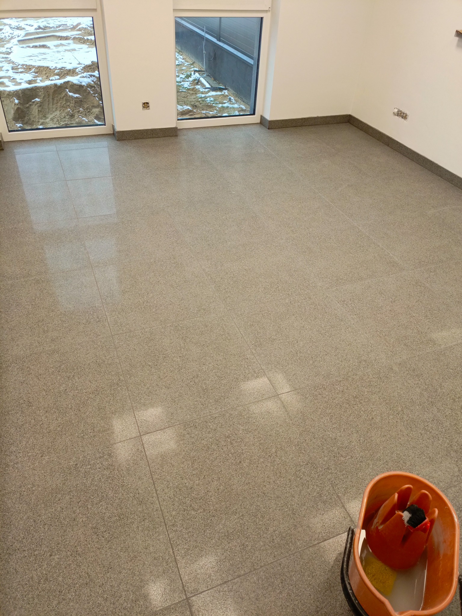 Wnętrze pomieszczenia z nową, jasną, gładką podłogą wyłożoną kwadratowymi płytkami. Widoczny kubeł z mopem w rogu, sugerujący świeżo zakończone prace porządkowe. Za oknami widoczny teren budowy.