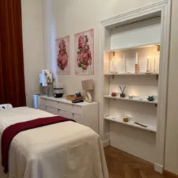 Przytulne wnętrze gabinetu z łóżkiem do zabiegów, białymi meblami, dekoracyjnymi świecami w półce i plakatami anatomicznymi na ścianie.